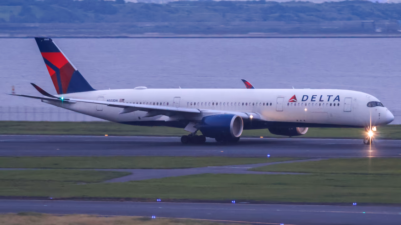 A Delta Air Lines Airbus A350 taxiing on a wet runway at dusk, its red and blue tail lit against grey water and low hills behind the airfield