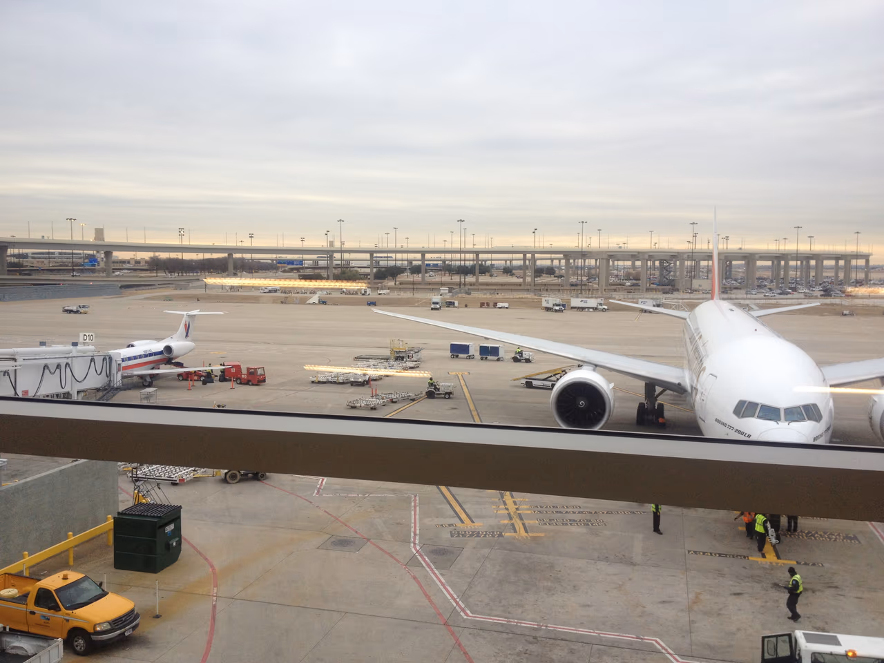 An American Airlines Boeing 777-200LR parked at a gate seen through a terminal window at Dallas Fort Worth, with a regional jet at the next stand, ground crew and baggage carts on the apron and an elevated roadway beyond