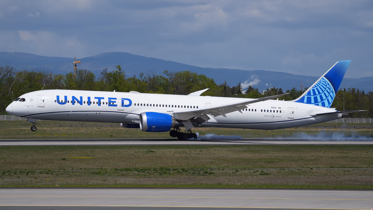A United Airlines Boeing 787-10 Dreamliner touching down on a runway with tire smoke at the main gear, wooded hills and a grey sky behind the airfield