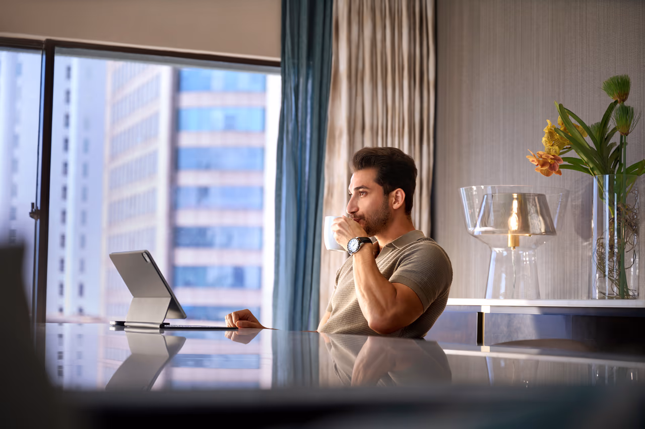 Man sitting at a table drinking from a mug while looking at a tablet in a suite at Hilton Colombo