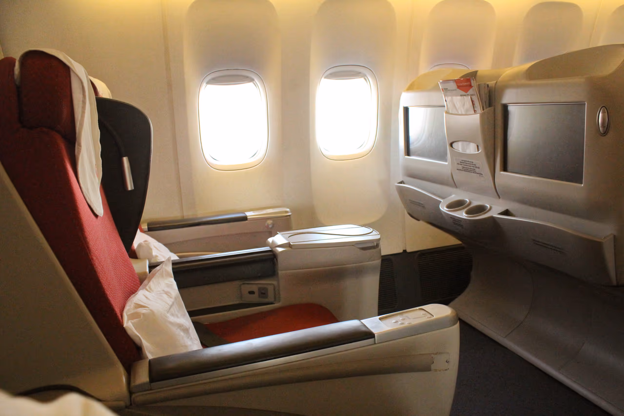 A widebody business class cabin with red lie-flat seats, seatback screens and white pillows, photographed from the aisle beside two cabin windows