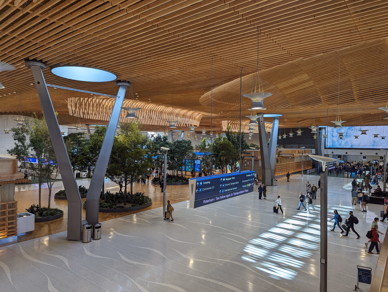 The undulating mass timber roof and indoor trees of Portland International Airport's main terminal hall, with travelers crossing the floor below a blue wayfinding sign