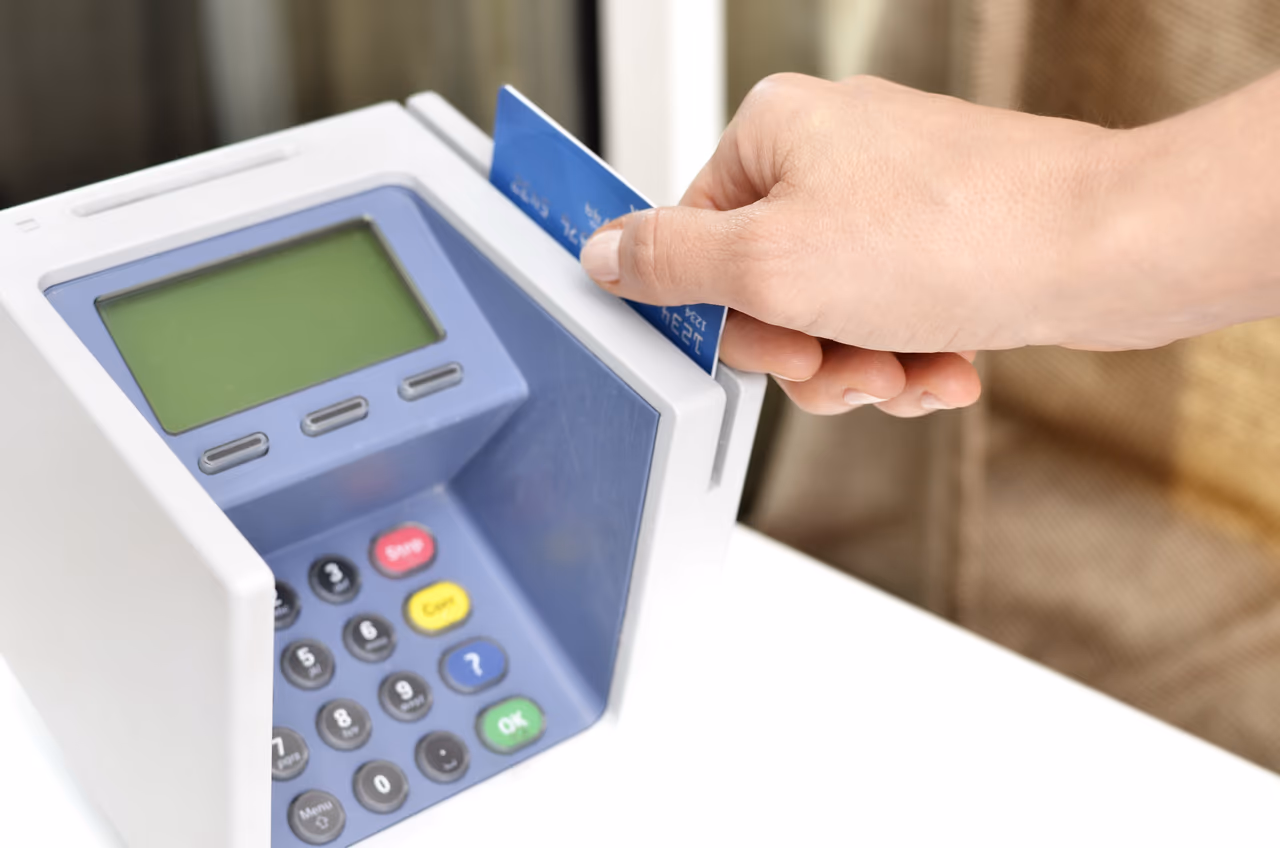 A hand inserting a blue credit card into a countertop card payment terminal with a numeric keypad and a green display