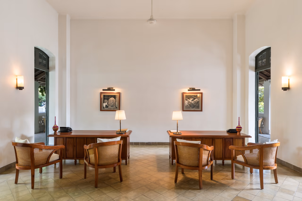 Two polished wooden reception desks with table lamps and cane-backed armchairs in a white-walled hotel lobby with arched doorways and a tiled floor