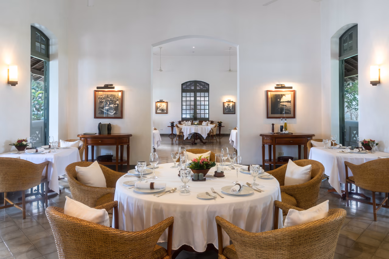 A restaurant dining room with white-clothed round tables set with glassware, wicker armchairs and framed black and white photographs on white walls