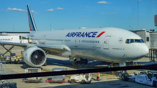 Air France Boeing 777 at a terminal gate with ground service vehicles, seen through an airport window