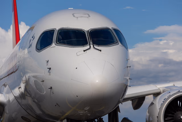 The nose of an Airbus A220 jet against a blue sky with clouds