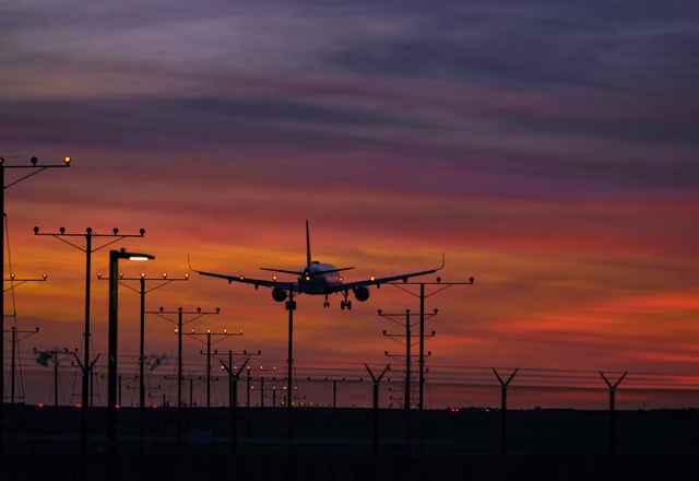 An airliner with its landing gear down above rows of runway approach lights, under a red, orange and purple sky