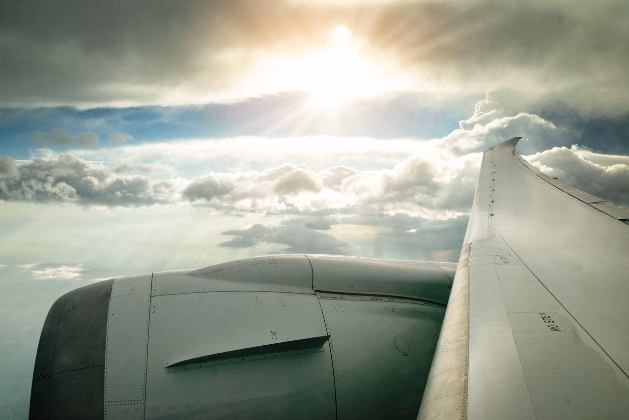 A jet engine and wing seen from a passenger window, with sunlight breaking through a layer of clouds