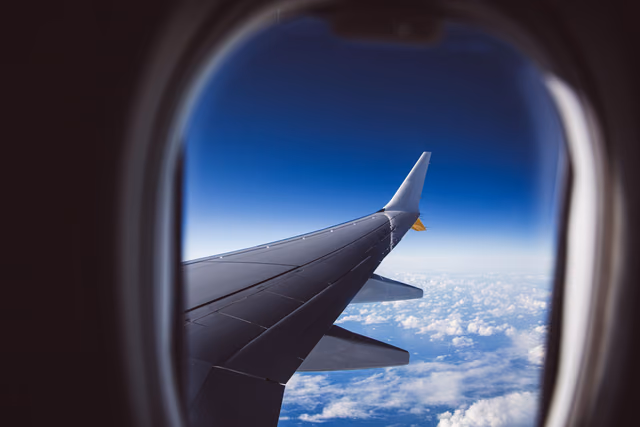 A gray airplane wing and winglet seen through an oval cabin window, above scattered white clouds and a deep blue sky