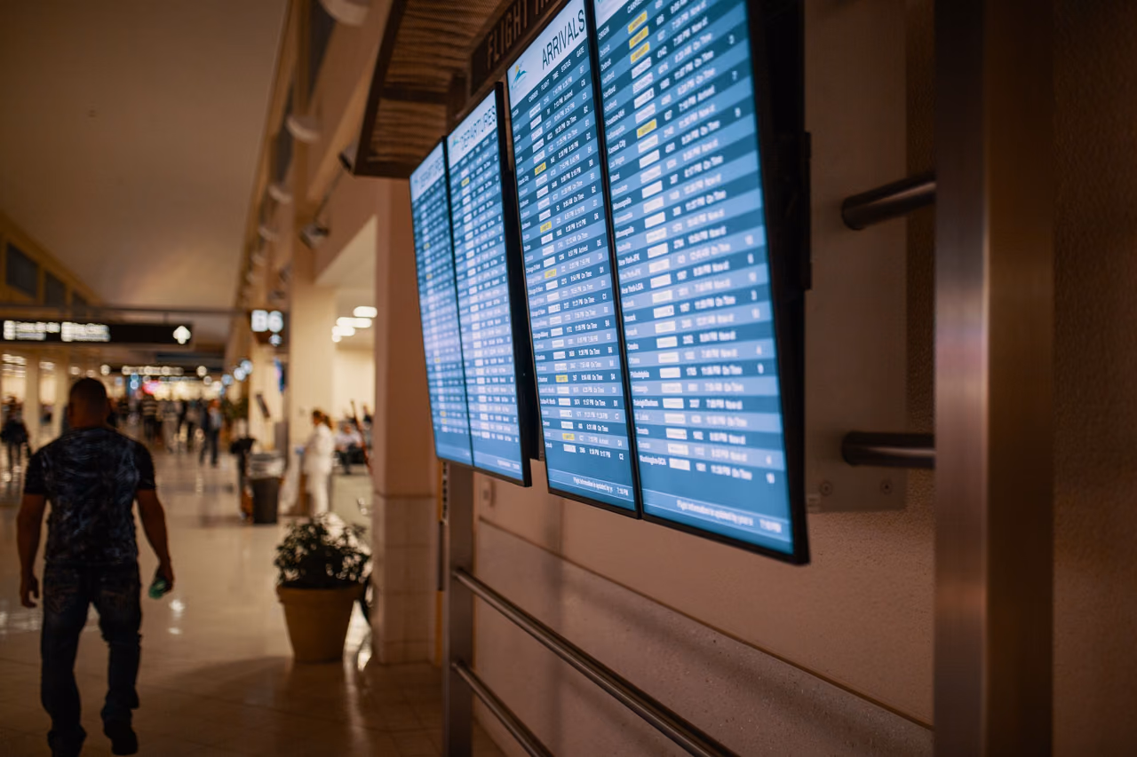 Flight information screens, one headed Arrivals, mounted on a wall in an airport terminal, with a man walking down the concourse