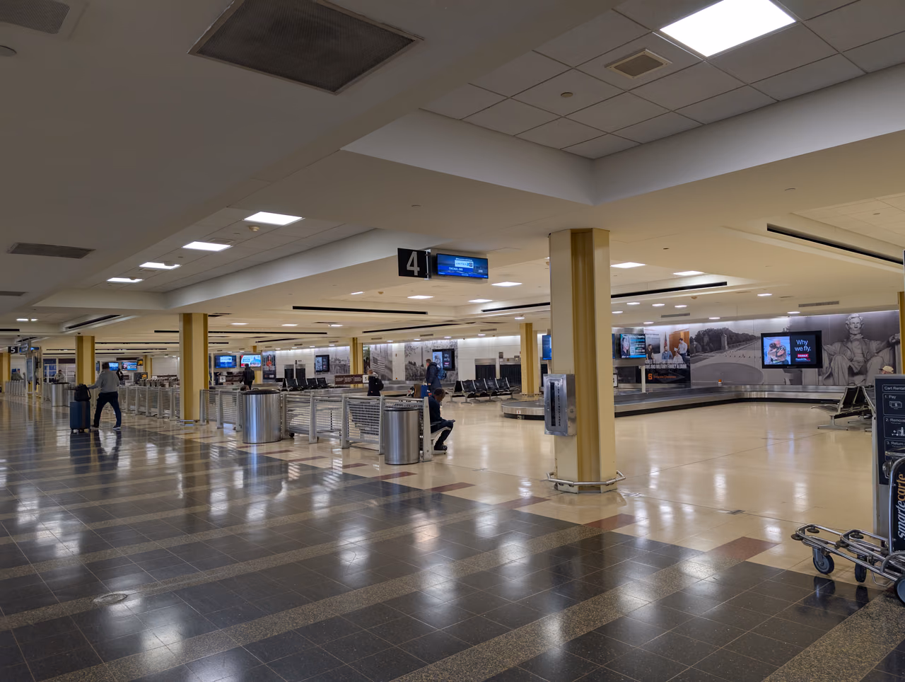 An airport baggage claim hall with an empty carousel, a sign reading 4 above it and a few travelers waiting near the belt