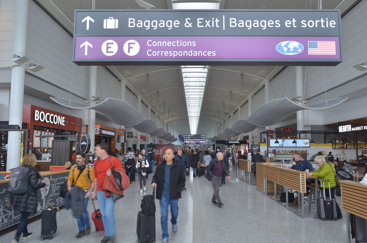 Airport concourse with an overhead sign pointing to Baggage & Exit and to Connections, travelers with rolling suitcases walking below
