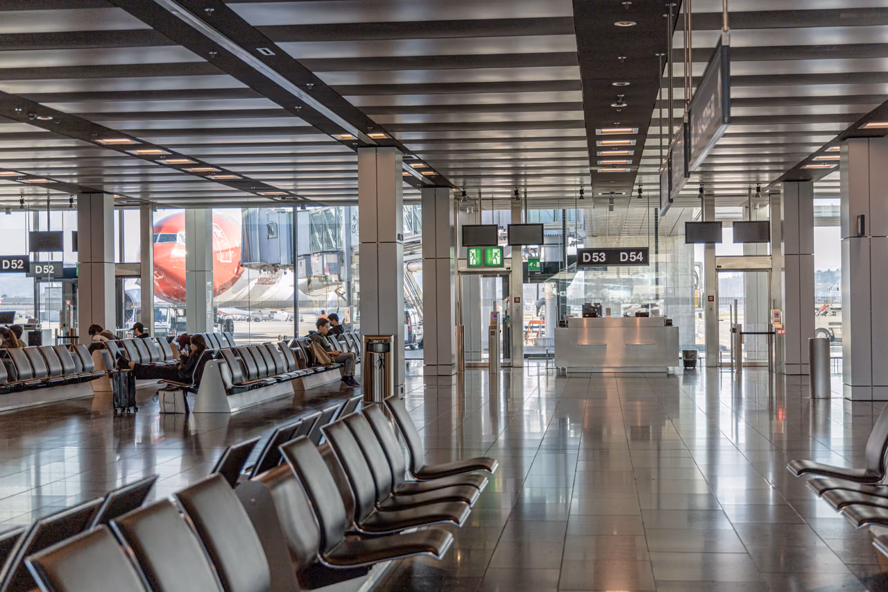 Rows of black leather seats in a bright airport departure area, with a jet parked at the gate beyond the windows
