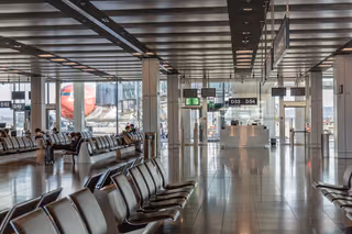 Rows of black leather seats in a bright airport departure area, with a jet parked at the gate beyond the windows