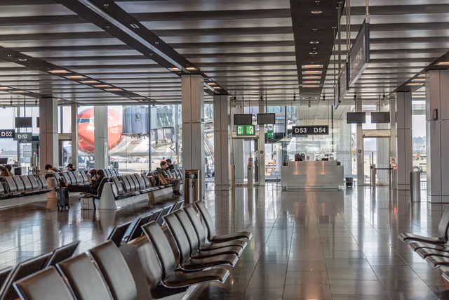 Rows of black leather seats in a bright airport departure area, with a jet parked at the gate beyond the windows