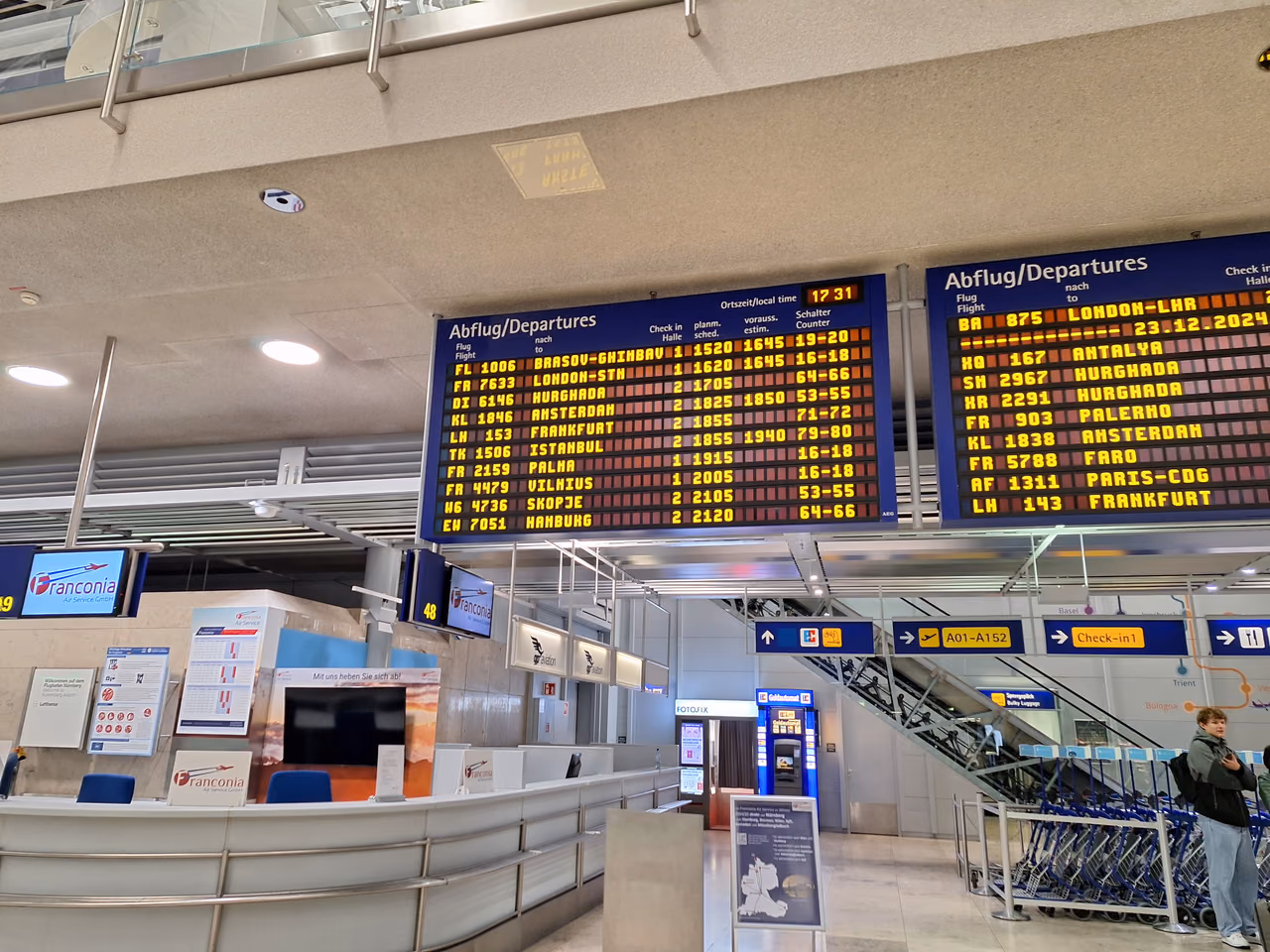 Two large blue departure boards above an airport concourse listing flights to European cities, with check-in signs and luggage carts below