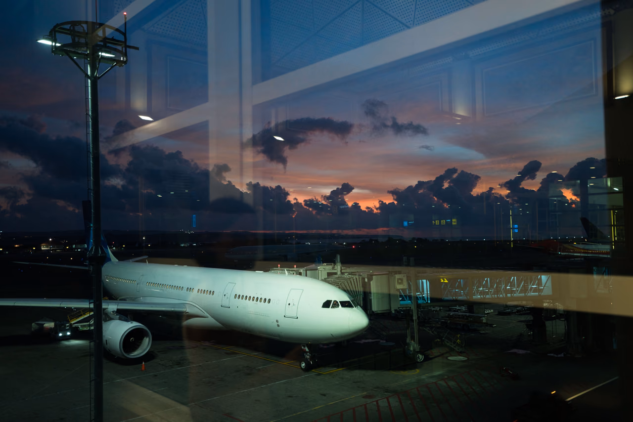 A white widebody jet parked at a jet bridge, seen through a terminal window at dusk under a pink and blue sky with dark clouds.