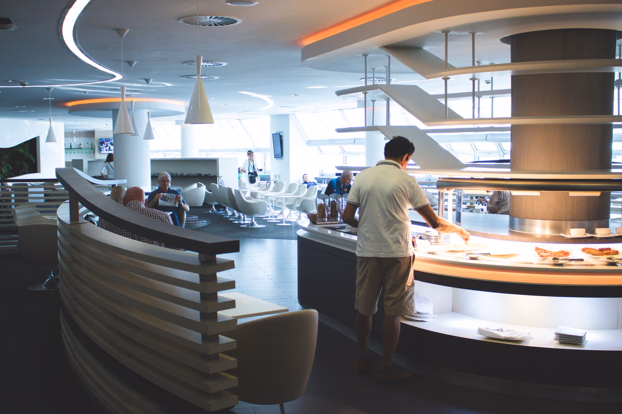 A traveler serves himself at a lit buffet counter in a bright airport lounge, with white swivel chairs and guests reading newspapers in the background.
