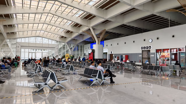 Travelers seated on steel chairs in a bright airport hall with a glass-paneled roof and a polished floor
