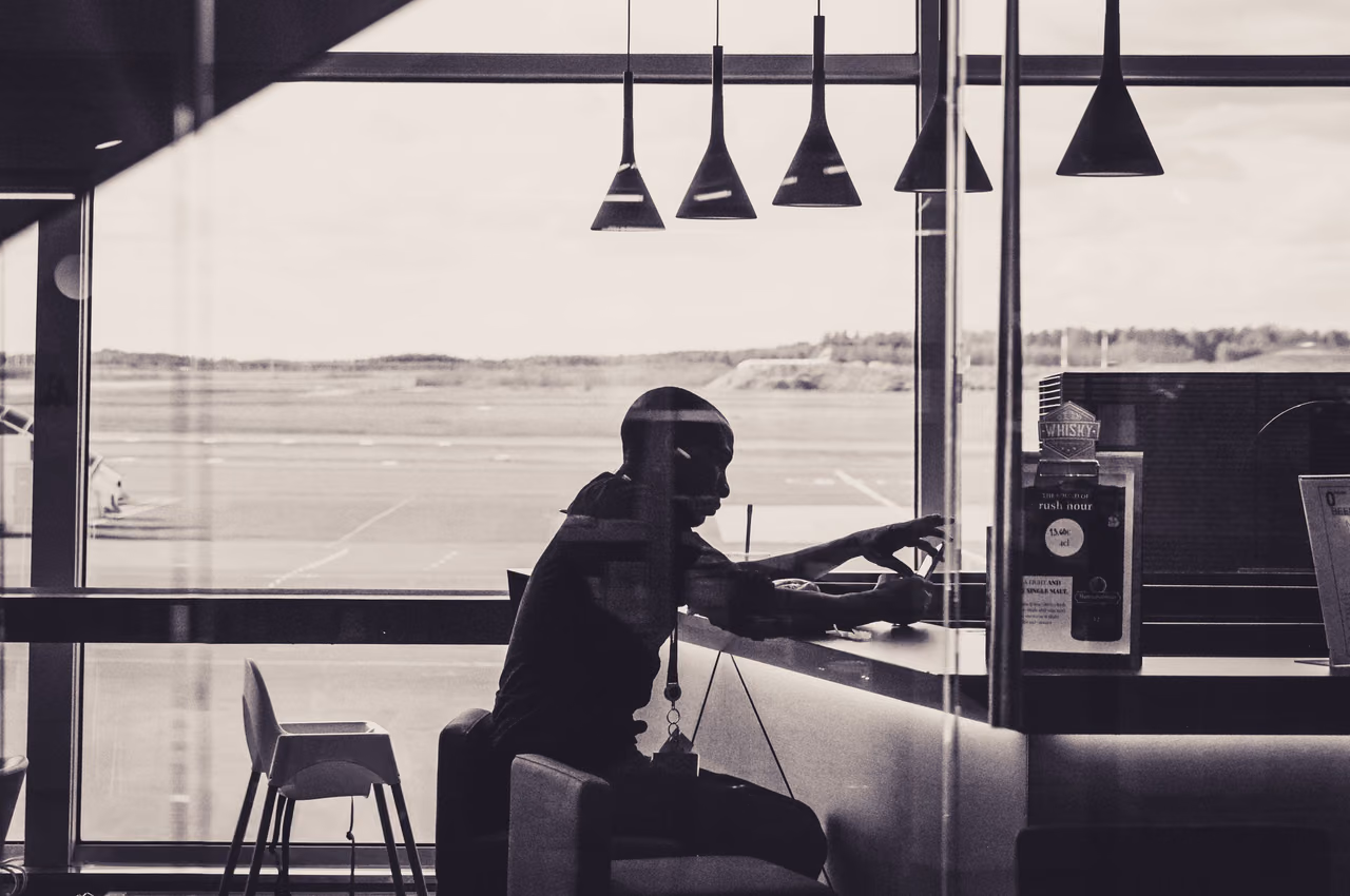 A black-and-white silhouette of a traveler seated at a counter under pendant lamps in an airport lounge, with the airfield visible through tall windows.