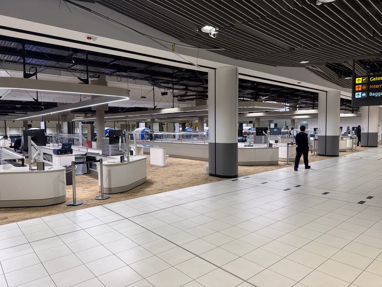 A wide airport security screening hall with empty X-ray belts, divesting benches and stacked trays, one officer standing near the lanes