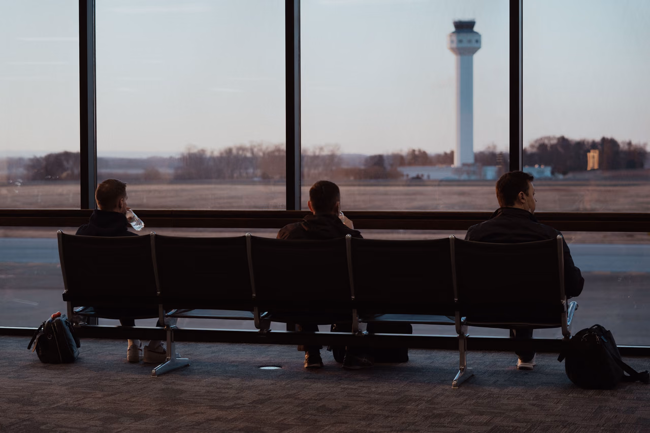 Three travelers sit on a row of airport terminal seats facing floor-to-ceiling windows that look out on the airfield and a control tower at dusk.