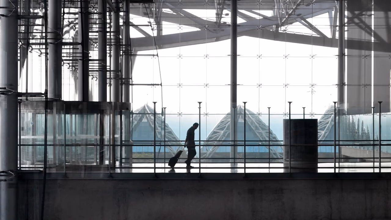 A traveler pulling a suitcase is silhouetted against the glass wall of a modern steel airport terminal.