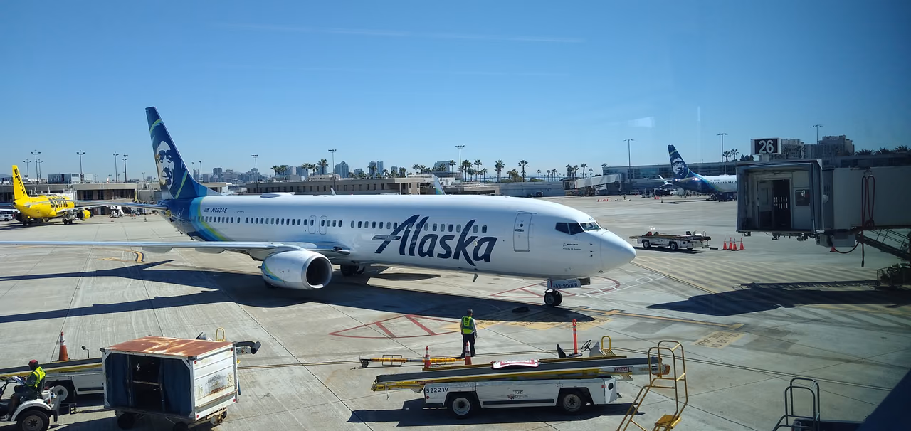 Alaska Airlines Boeing 737-900ER in white livery with a blue and teal tail bearing a portrait of an Alaska Native man, pulling onto a stand at San Diego International Airport as a ground crew member in a hi-vis vest guides it in
