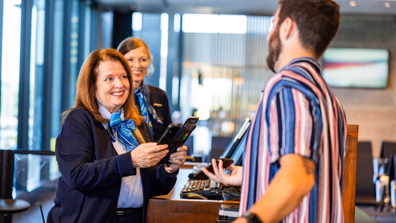 An Alaska Airlines gate agent smiles while scanning a passenger's phone boarding pass at the podium, with a second agent behind her