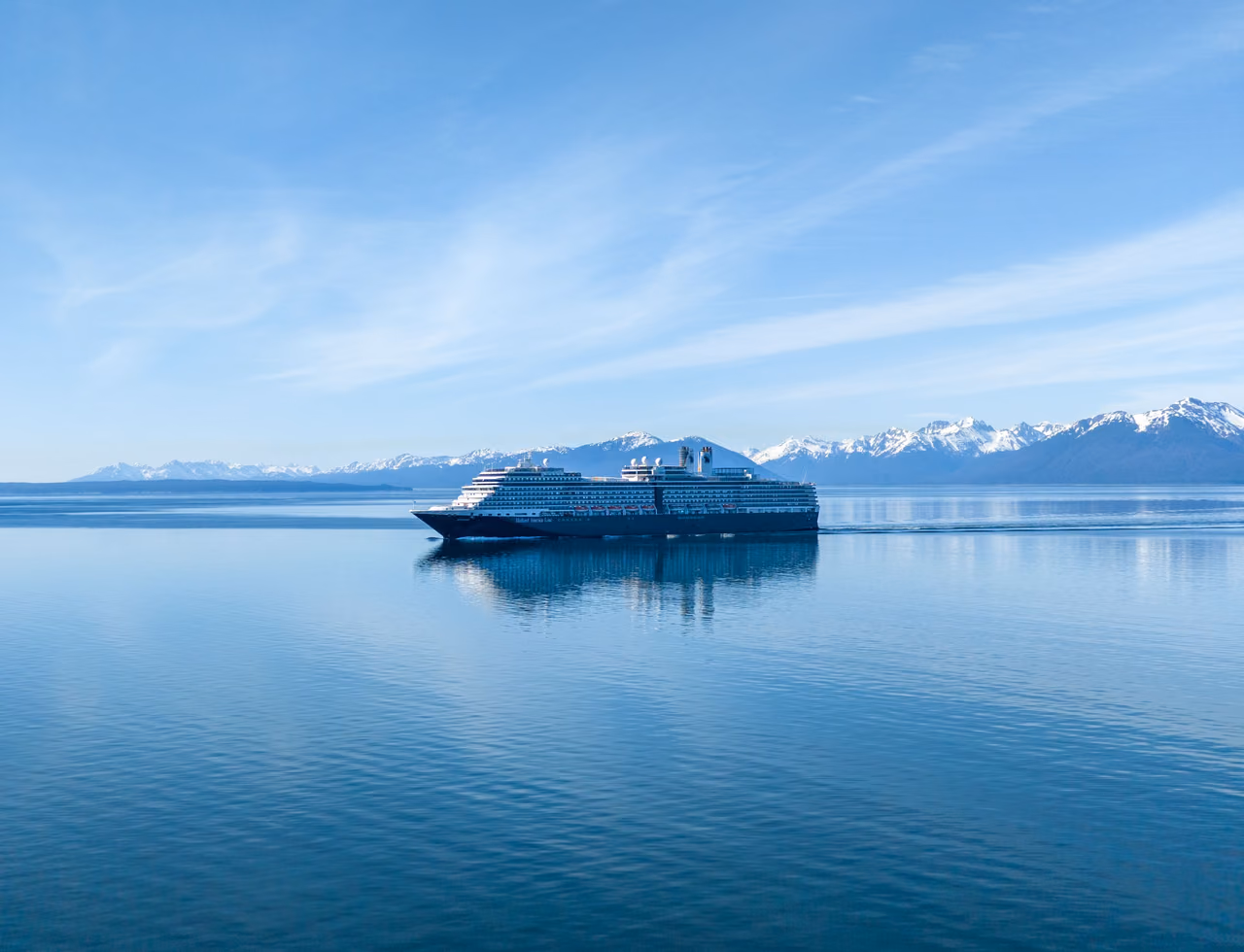 A large cruise ship crossing calm blue water in front of snow-capped mountains near Hoonah, Alaska