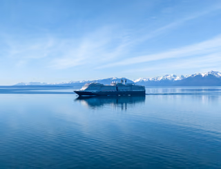 A large cruise ship crossing calm blue water in front of snow-capped mountains near Hoonah, Alaska