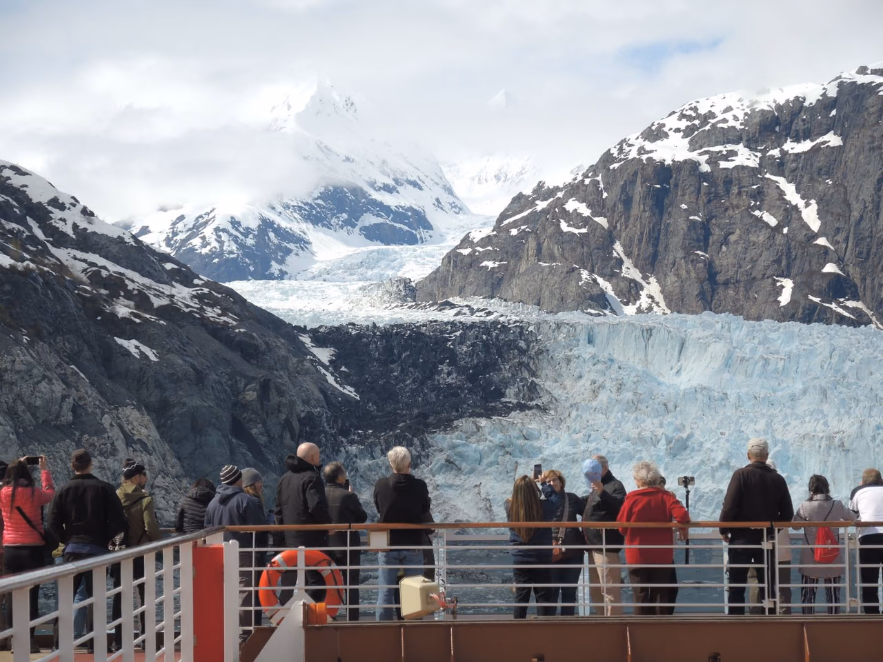 Passengers on a cruise ship deck looking at a glacier between snowy Alaska mountains