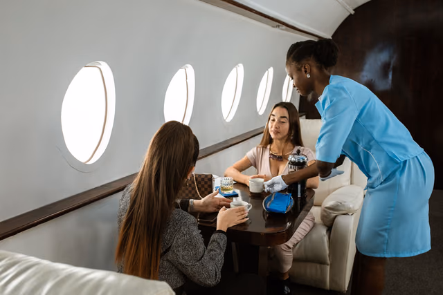 A flight attendant in a light blue uniform pouring coffee for two seated passengers at a table beside oval windows in a private jet cabin