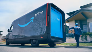 An Amazon Prime electric delivery van parked at the curb with its rear door open while a driver carries a package up the walk to a house