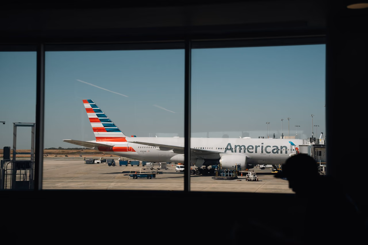 An American Airlines widebody jet at the gate, seen through a terminal window with a traveler in silhouette
