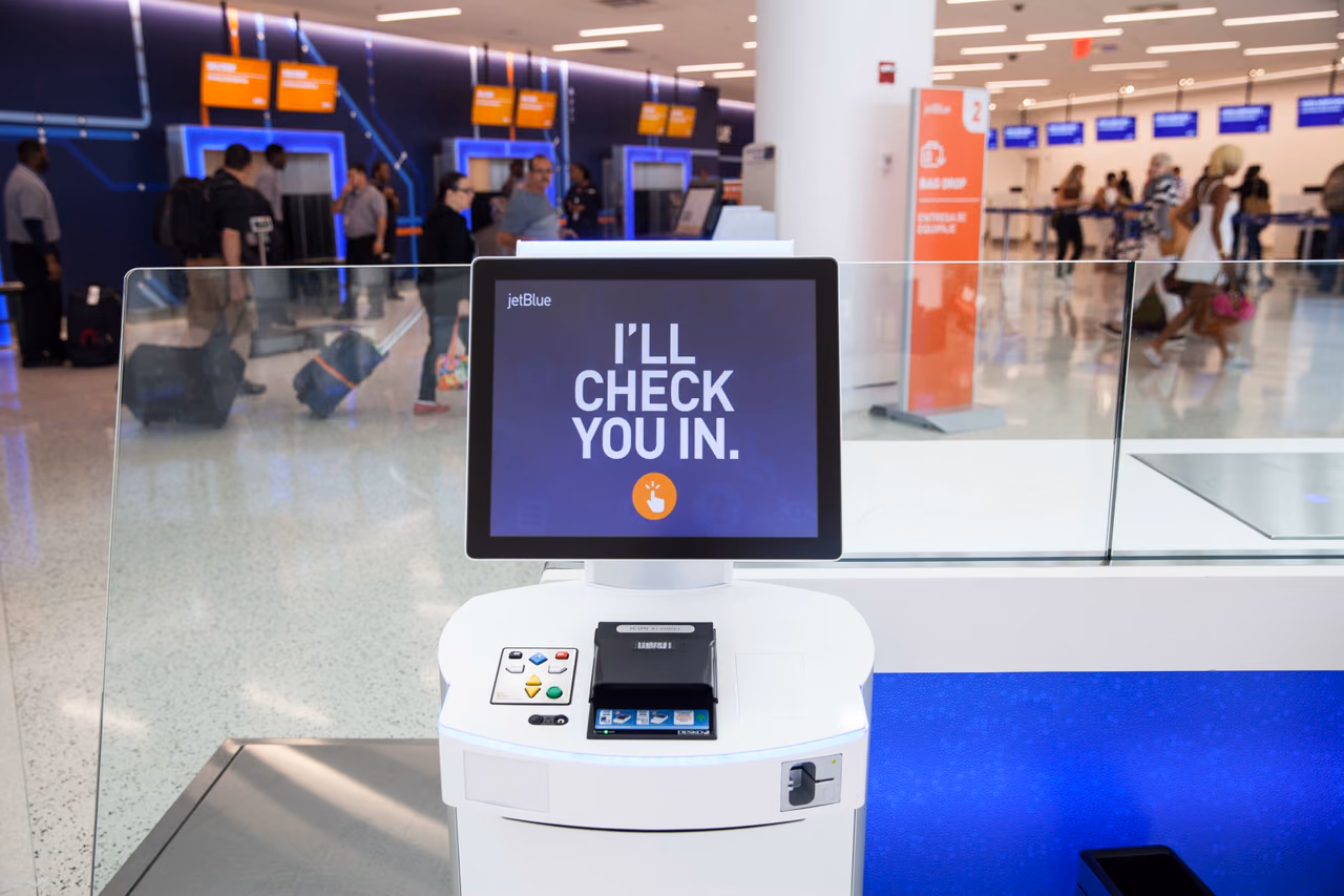A JetBlue self check-in kiosk with the screen message I'll check you in, in a terminal with travelers and bag drop counters behind it.