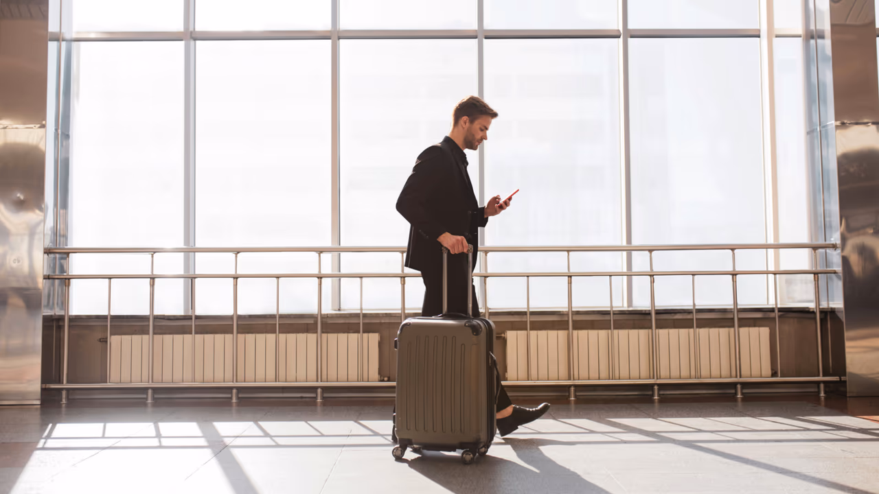 Business traveler walking through airport with suitcase while checking phone.