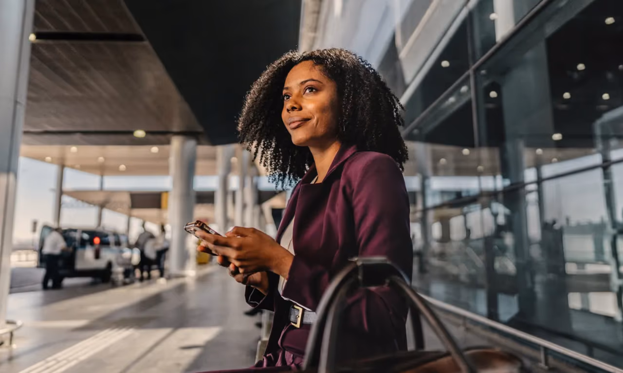 woman sitting on a bench holding her phone with her luggage sitting on the bench next to her