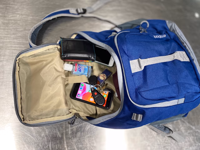 An open blue backpack on a metal table with two smartphones, a wallet, keys and hand sanitizer packed inside