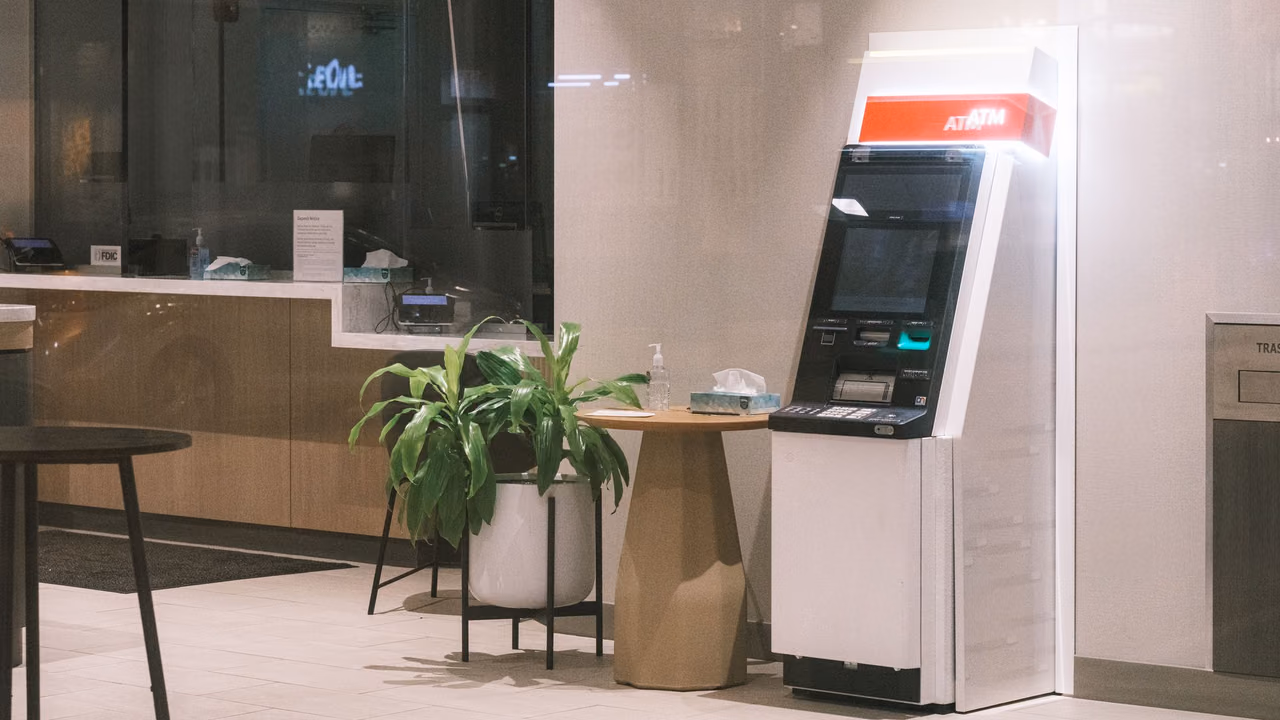 An ATM standing in a quiet, modern bank lobby next to a plant and a small table