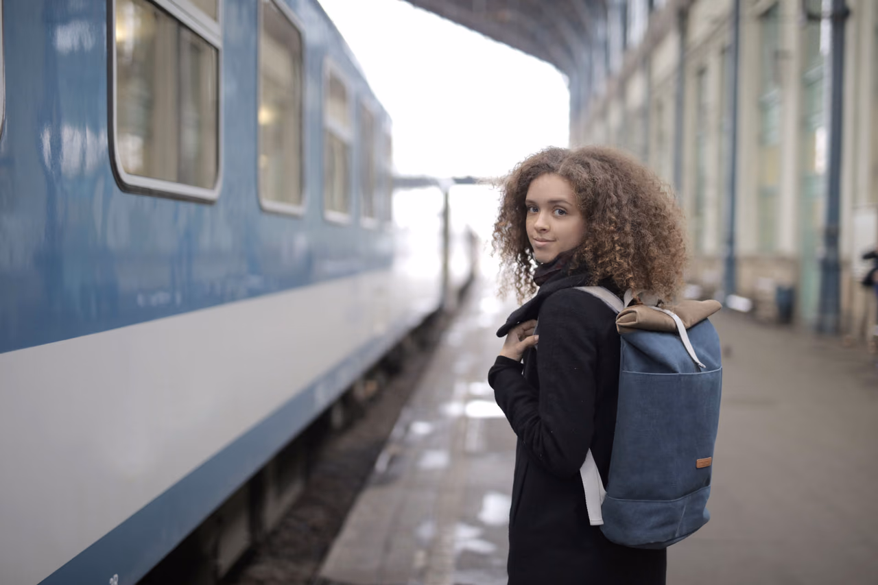 A young woman with a denim backpack stands on a covered train platform beside a blue passenger carriage, looking back over her shoulder.