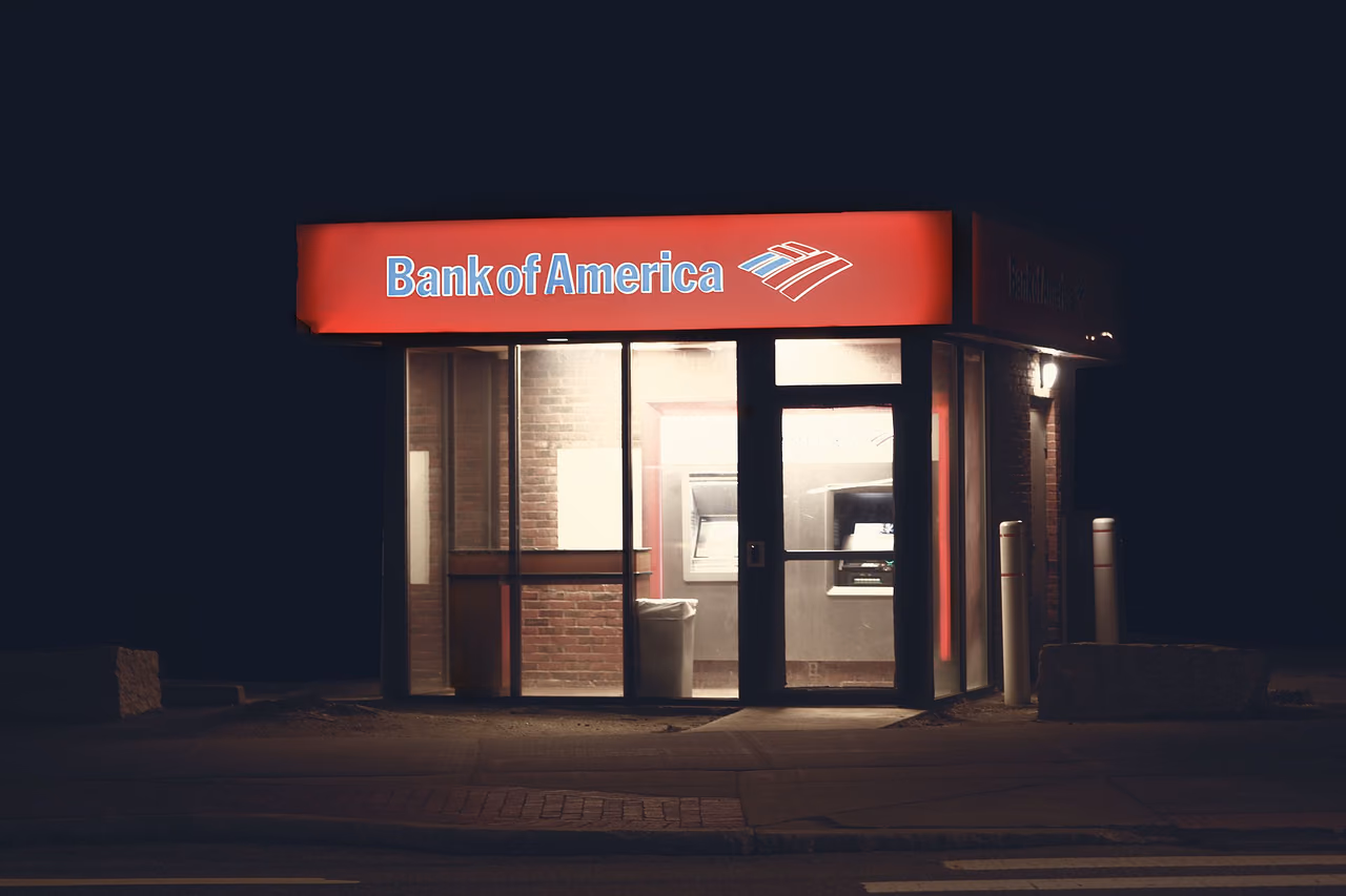 A lit Bank of America ATM vestibule with a red sign on a dark street at night