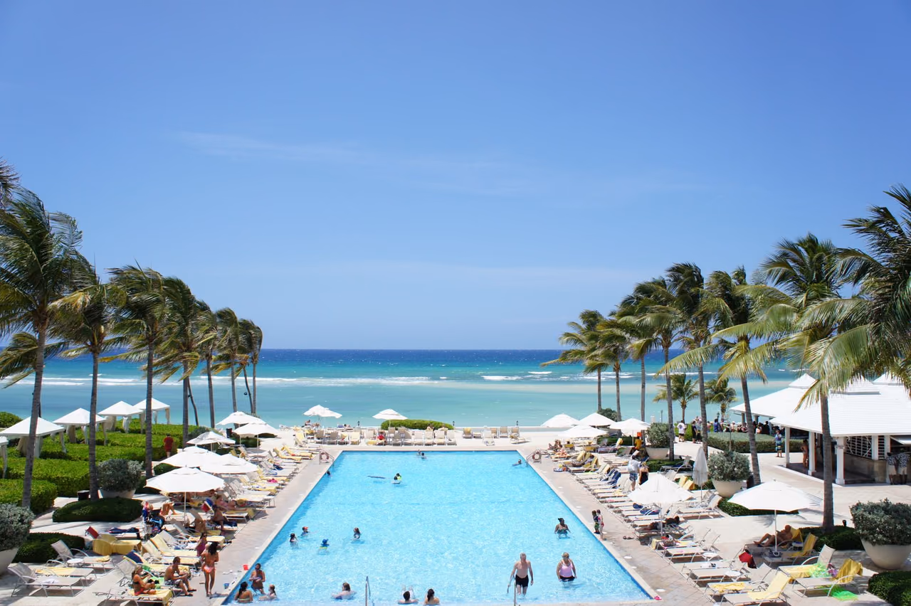 Beachfront resort pool lined with loungers and umbrellas, framed by palm trees and a turquoise sea