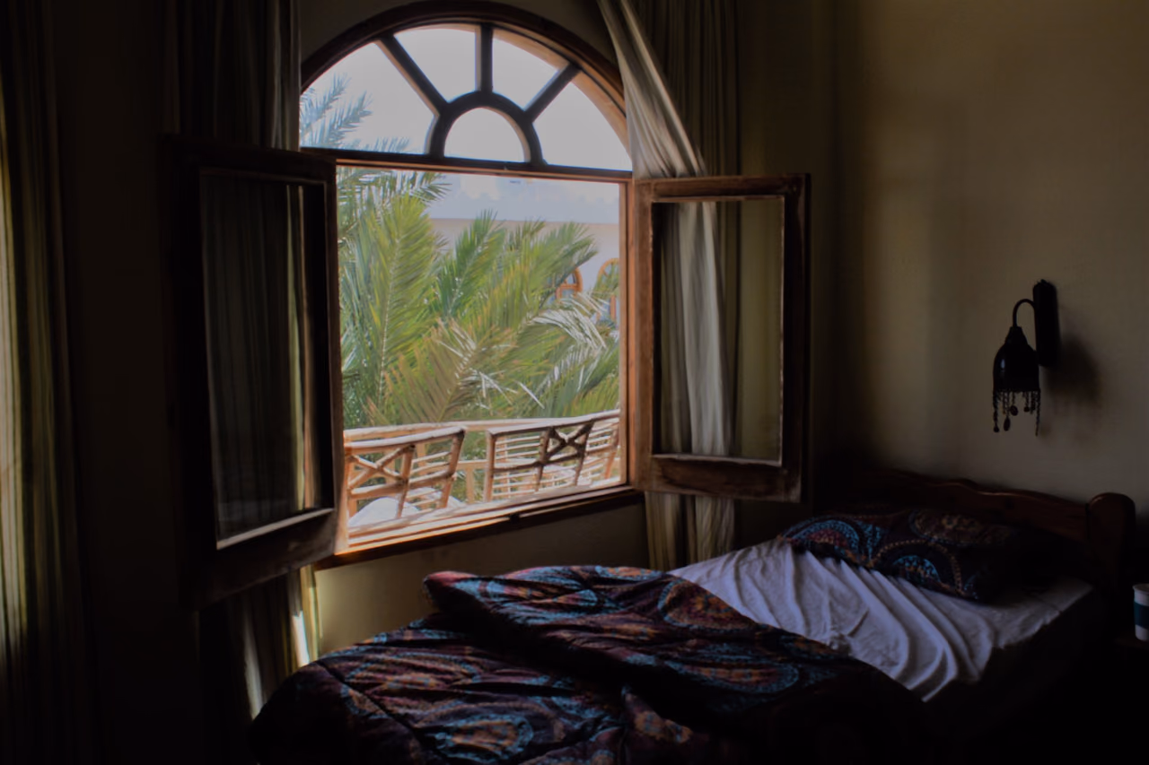 A bedroom with an open wooden window frame and a palm tree outside