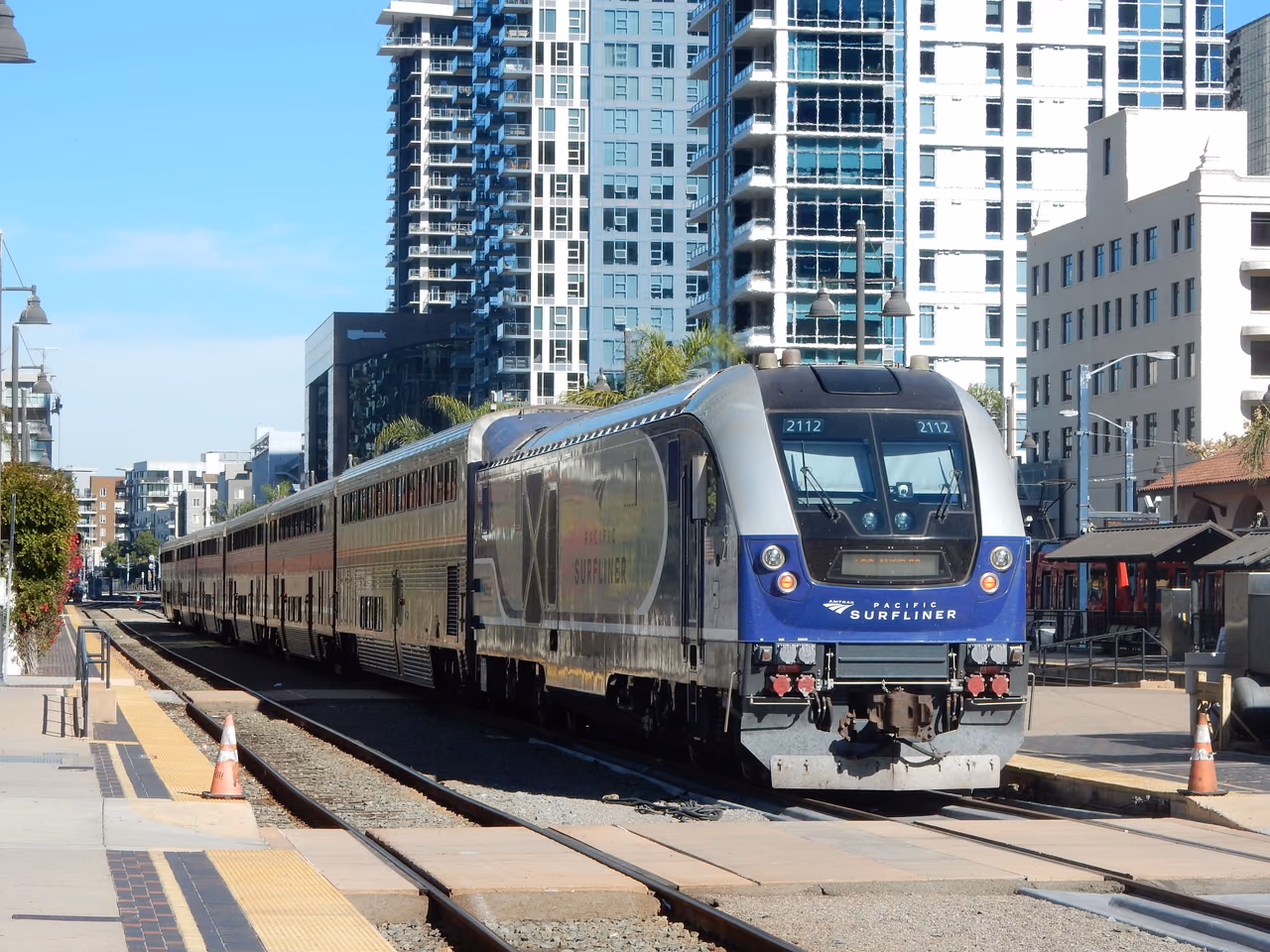An Amtrak Pacific Surfliner train with a blue and silver locomotive pulls into a downtown San Diego station platform beside high-rise buildings.
