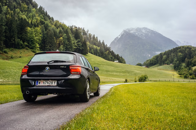 A black BMW hatchback seen from behind on a wet country road beside green meadows, with forested hills and a snow-capped mountain ahead