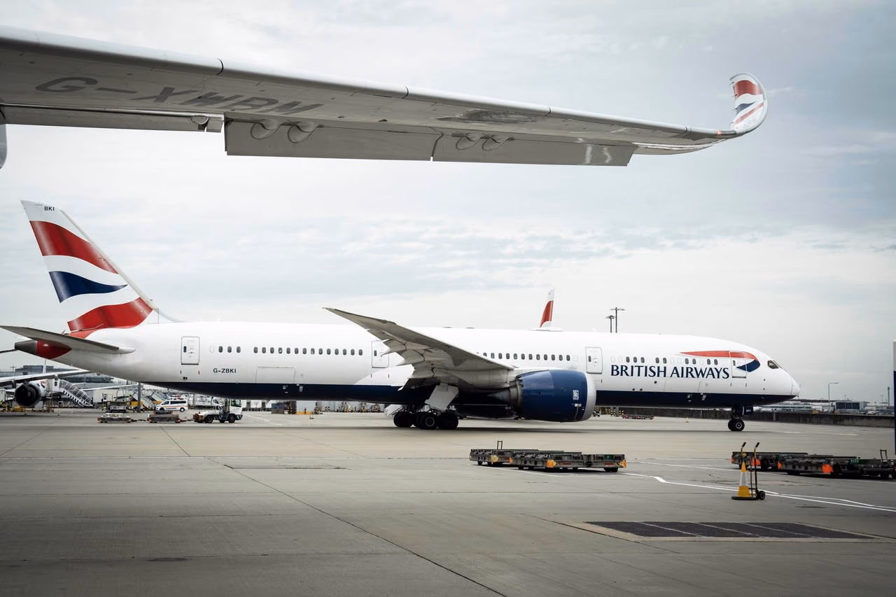 British Airways Boeing 787 Dreamliner parked on the apron at London Heathrow under an overcast sky, with another aircraft's wingtip overhead