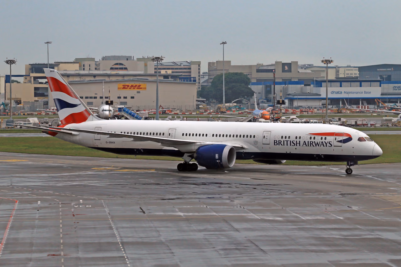 A British Airways twin-engine jet taxiing on a wet airport apron, with hangars and terminal buildings behind it
