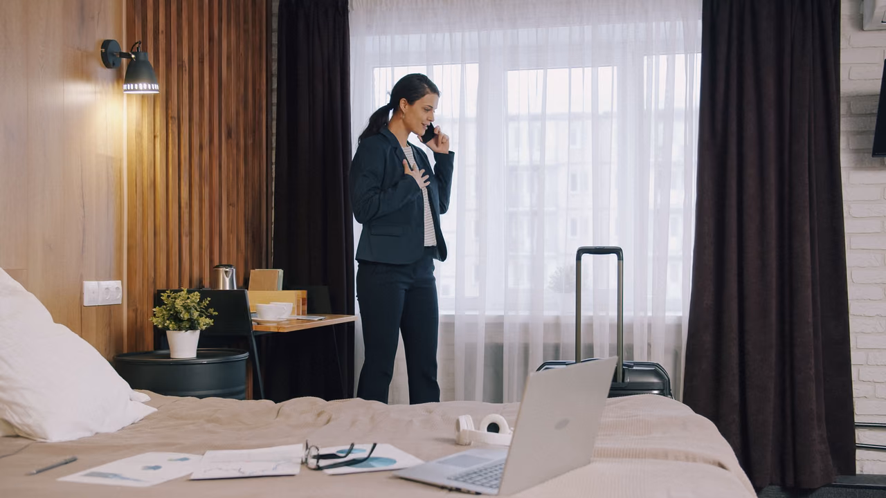 A woman in a dark suit takes a phone call by the window of a hotel room, with a laptop, papers and glasses on the bed and a suitcase behind her